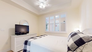 Un dormitorio con cama, televisor sobre una cómoda, ventilador de techo y ventana con persianas.
