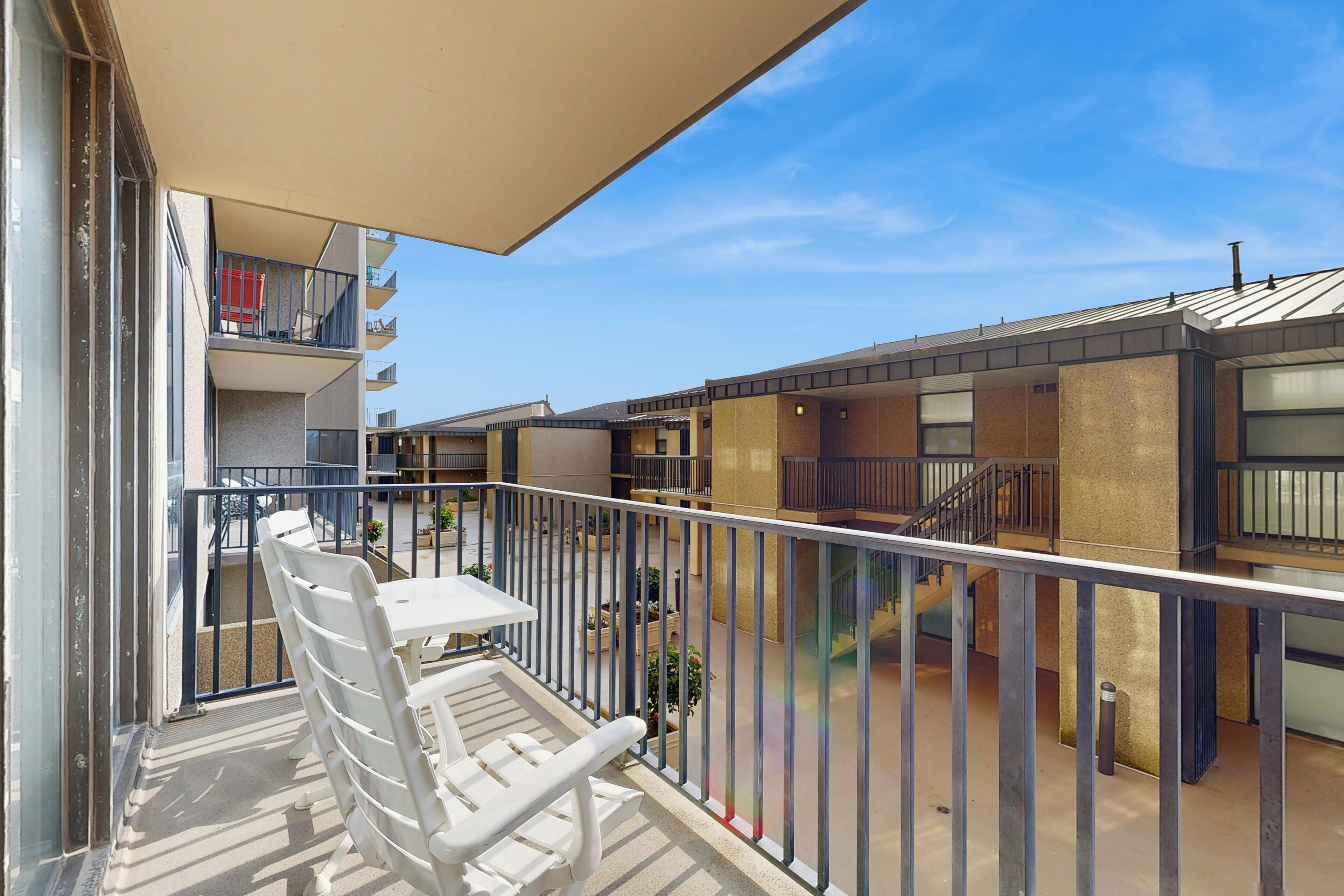 A balcony with white plastic chairs and a table, overlooking a courtyard with buildings and a clear sky.