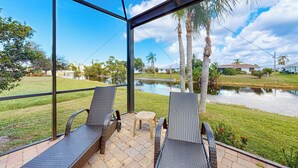 A covered patio area with two wicker chairs and a small stool, overlooking a pond and a grassy area with palm trees. - 4BR Home w/ Outdoor Patio & In-ground Pool (Rotonda West)