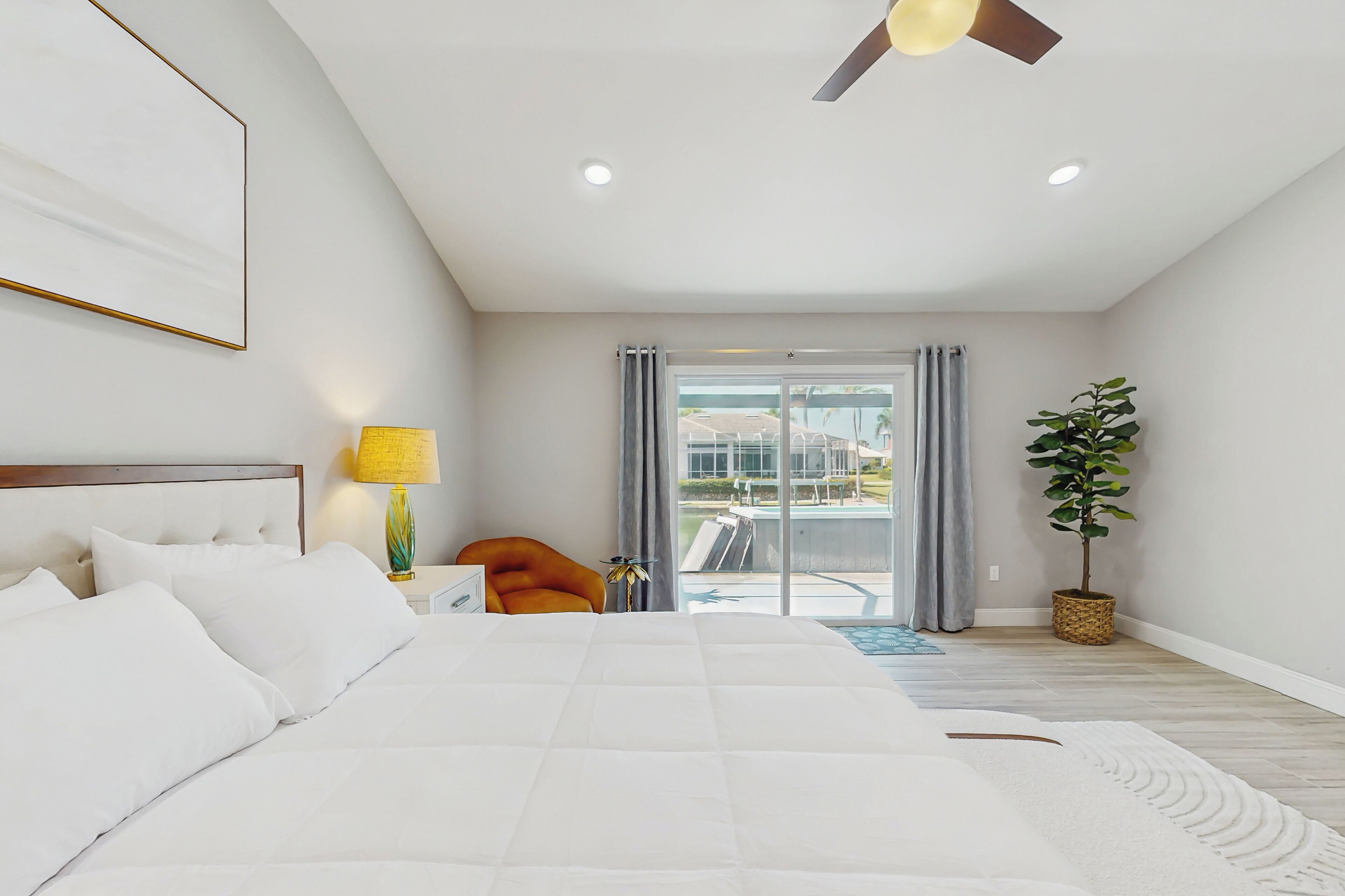 A modern hotel room with a large bed, a ceiling fan, a view of the outdoors through a sliding door, and a potted plant.