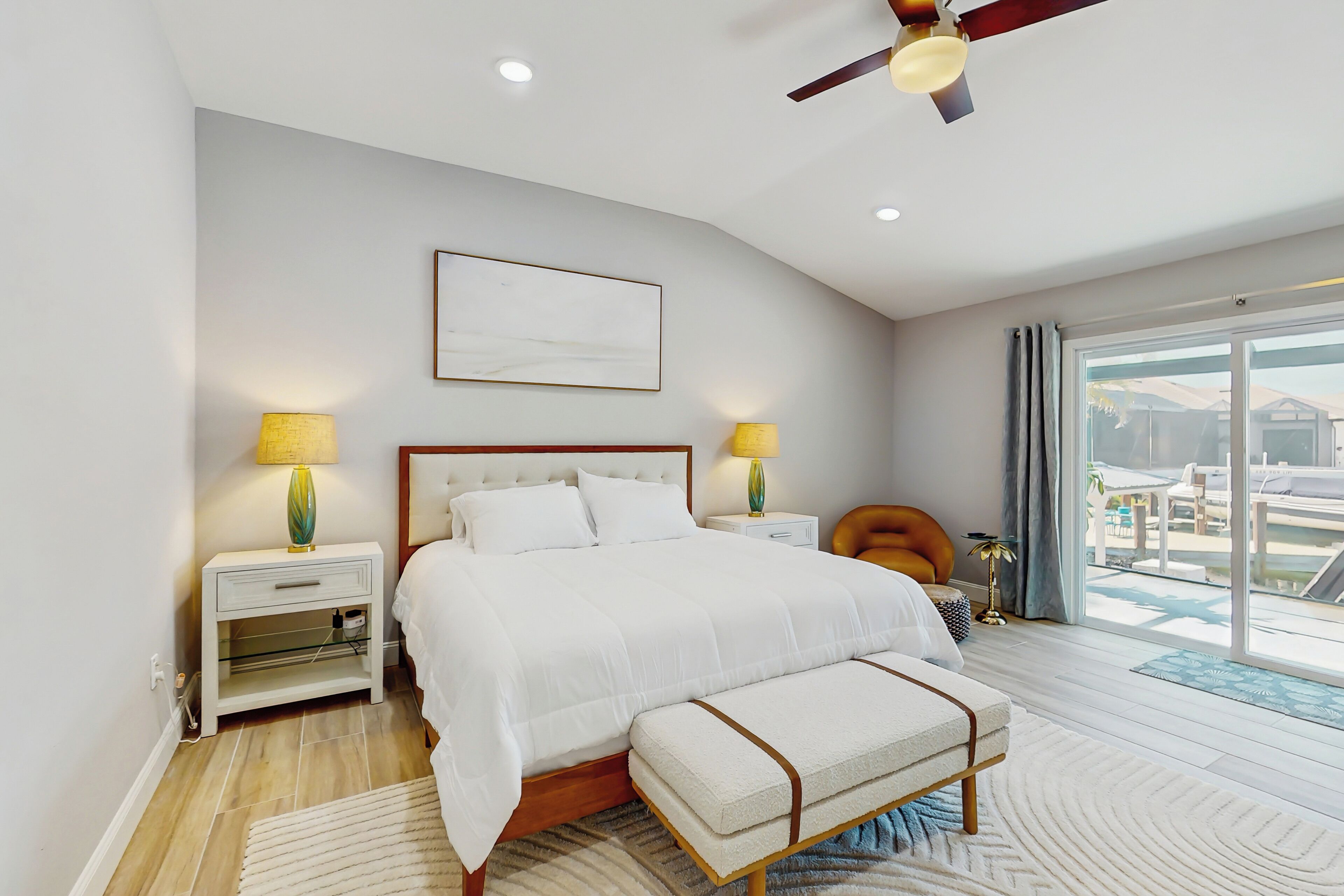 A modern bedroom with a large bed, bedside lamps, a ceiling fan, and a view of the outdoors.