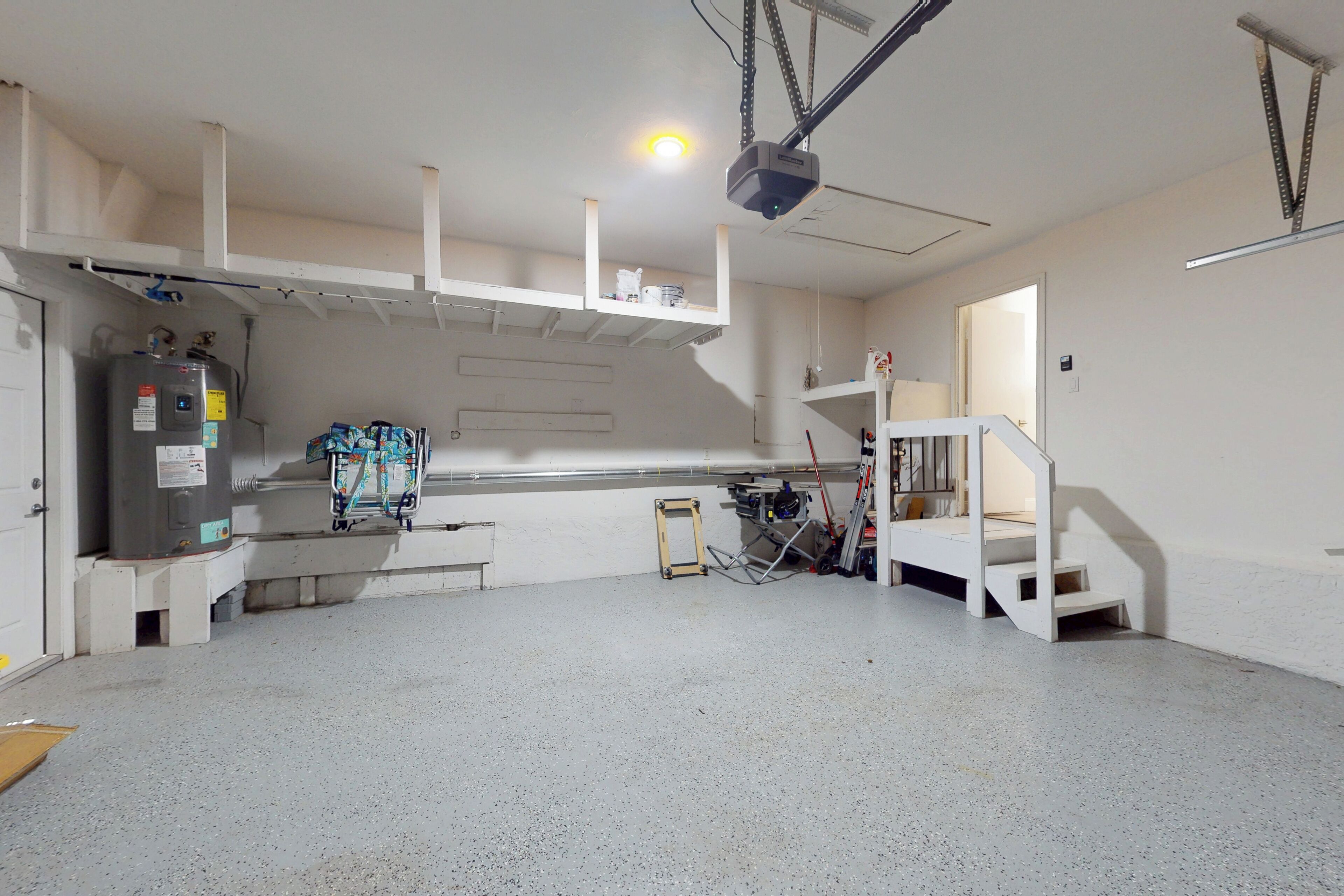 A spacious, well-lit industrial room with a large workbench, a water cooler, and a staircase leading to an upper level.