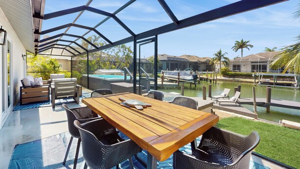 En overdækket terrasse med et træbord og stole, med udsigt over en swimmingpool og et anlagt område med palmer.