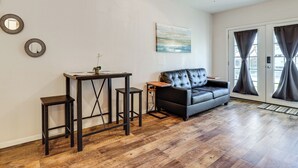 A living room with a black leather sofa, a wooden coffee table, and two black bar stools. There is a ceiling fan, a painting on the wall, and a large window with curtains. - 9 Mi to Rangers & Cowboys Home Games: DFW Studio (Grand Prairie)