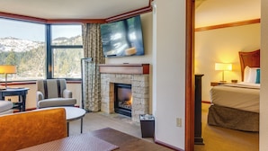 A hotel room with a stone fireplace, a television mounted on the wall, a bed, and a view of the mountains through large windows. - Everline Resort Suite: North Lake Tahoe Luxury! (Olympic Valley)