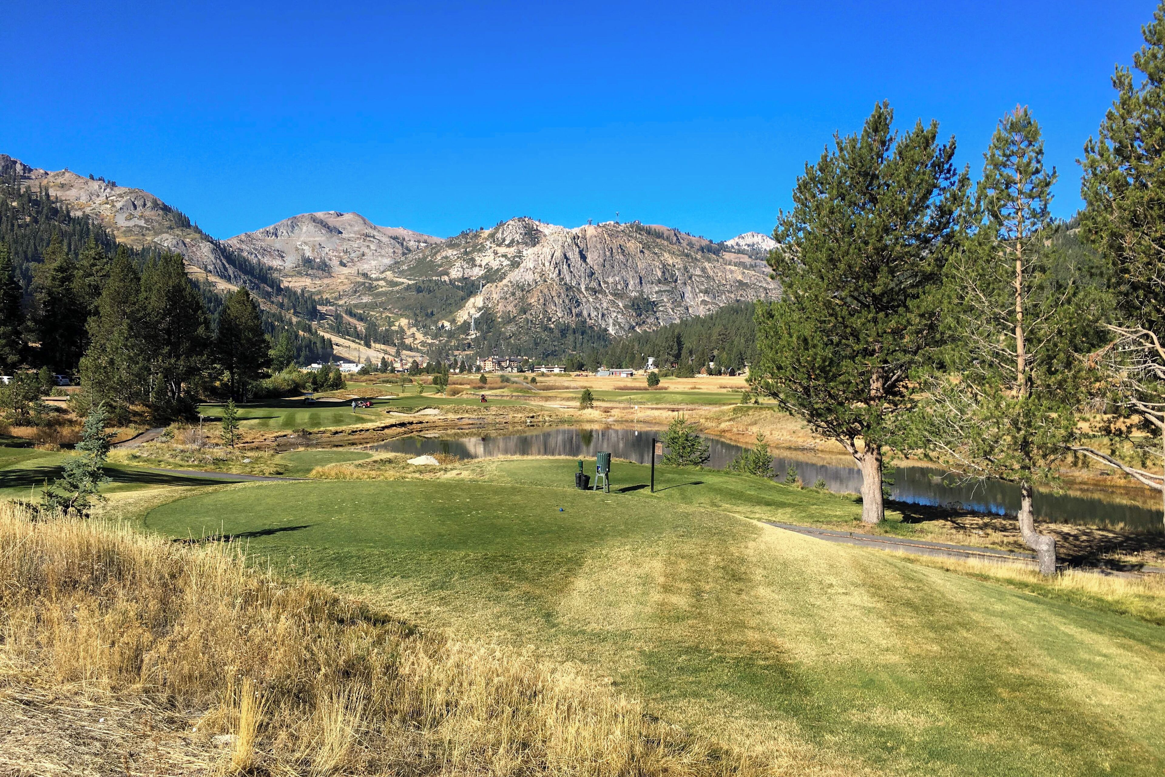 Un campo de golf con un estanque, montañas al fondo y un cielo azul despejado.