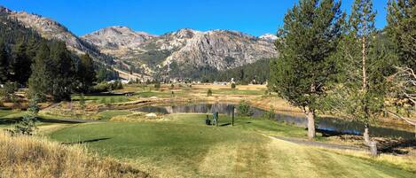 Un campo de golf con un estanque, montañas al fondo y un cielo azul despejado.