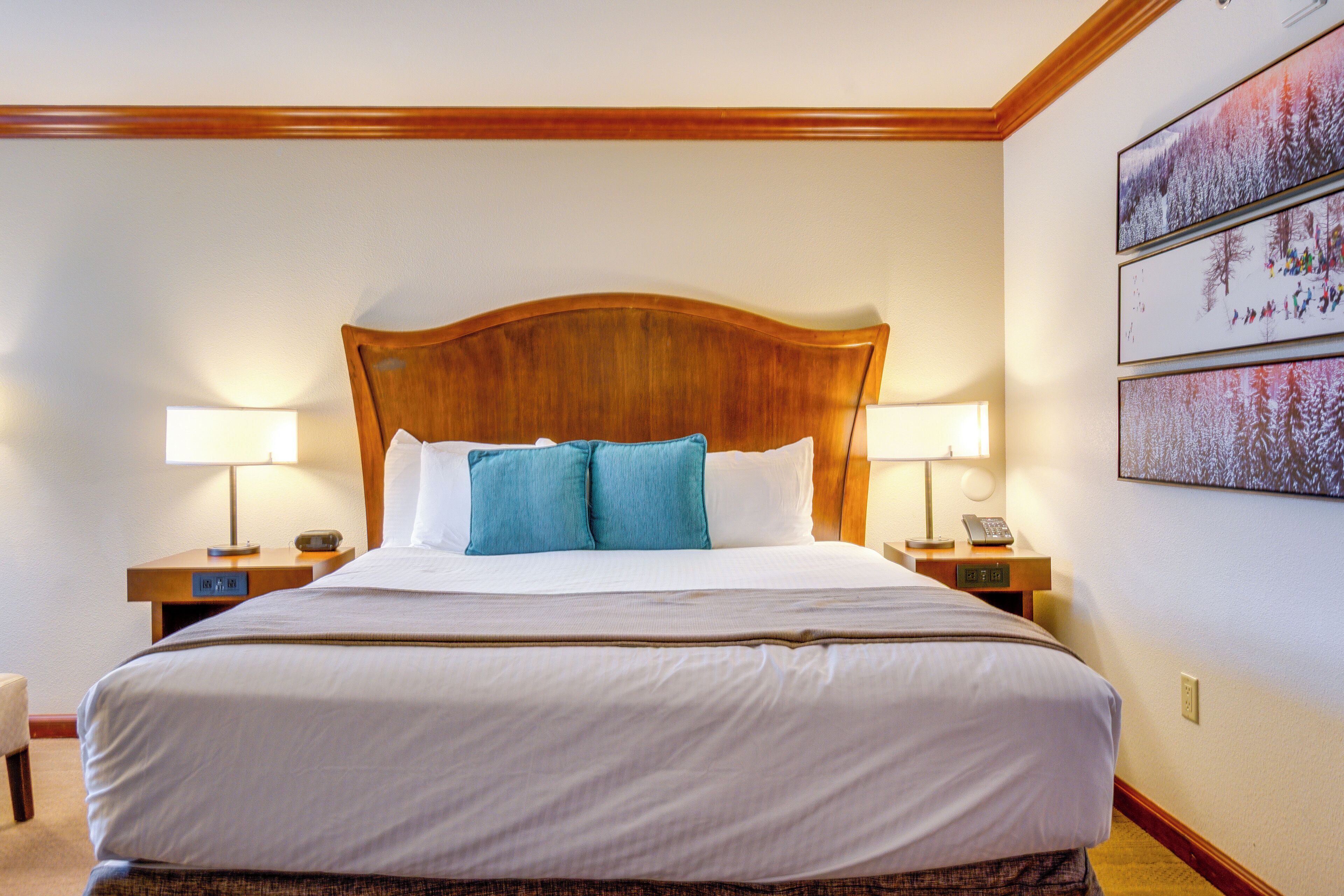 A neatly made bed with white and blue bedding, wooden headboard, two bedside tables with lamps, and a wall with framed pictures.