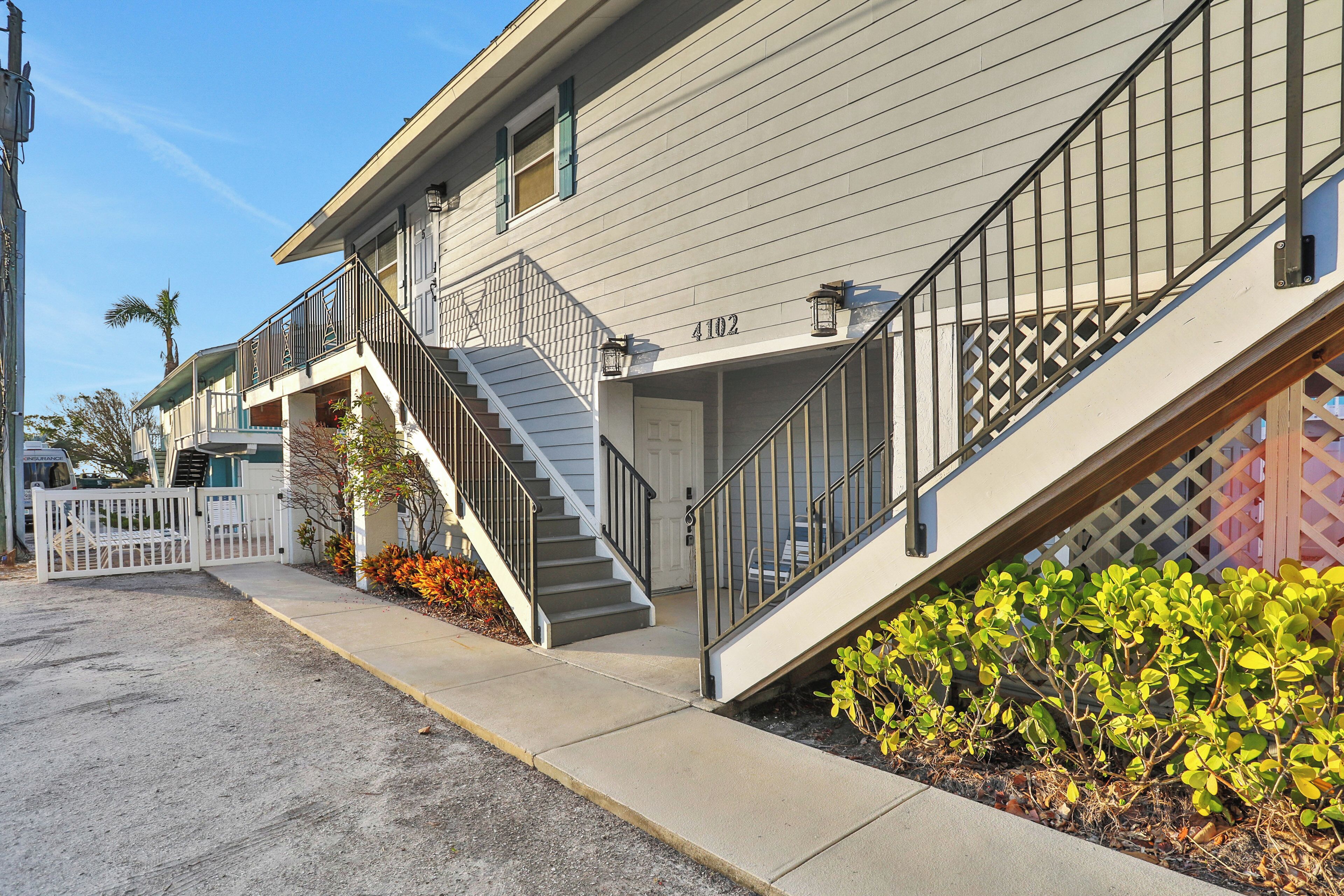 A two-story building with a staircase leading to the entrance, a gated driveway, and a small garden area with shrubs.