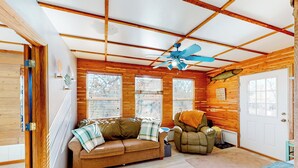 Une pièce chaleureuse avec des murs en bois, un ventilateur de plafond, un canapé en cuir marron, un fauteuil vert et une porte blanche.