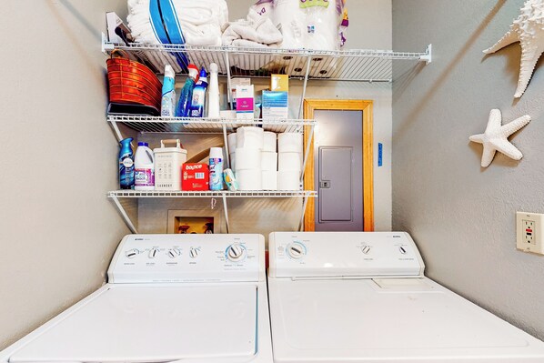 A laundry area with two front-loading washing machines, shelves above containing various laundry supplies, and a door leading to another room. - Relax at 'the Cove' Waterfront Getaway (Eagle Rock)