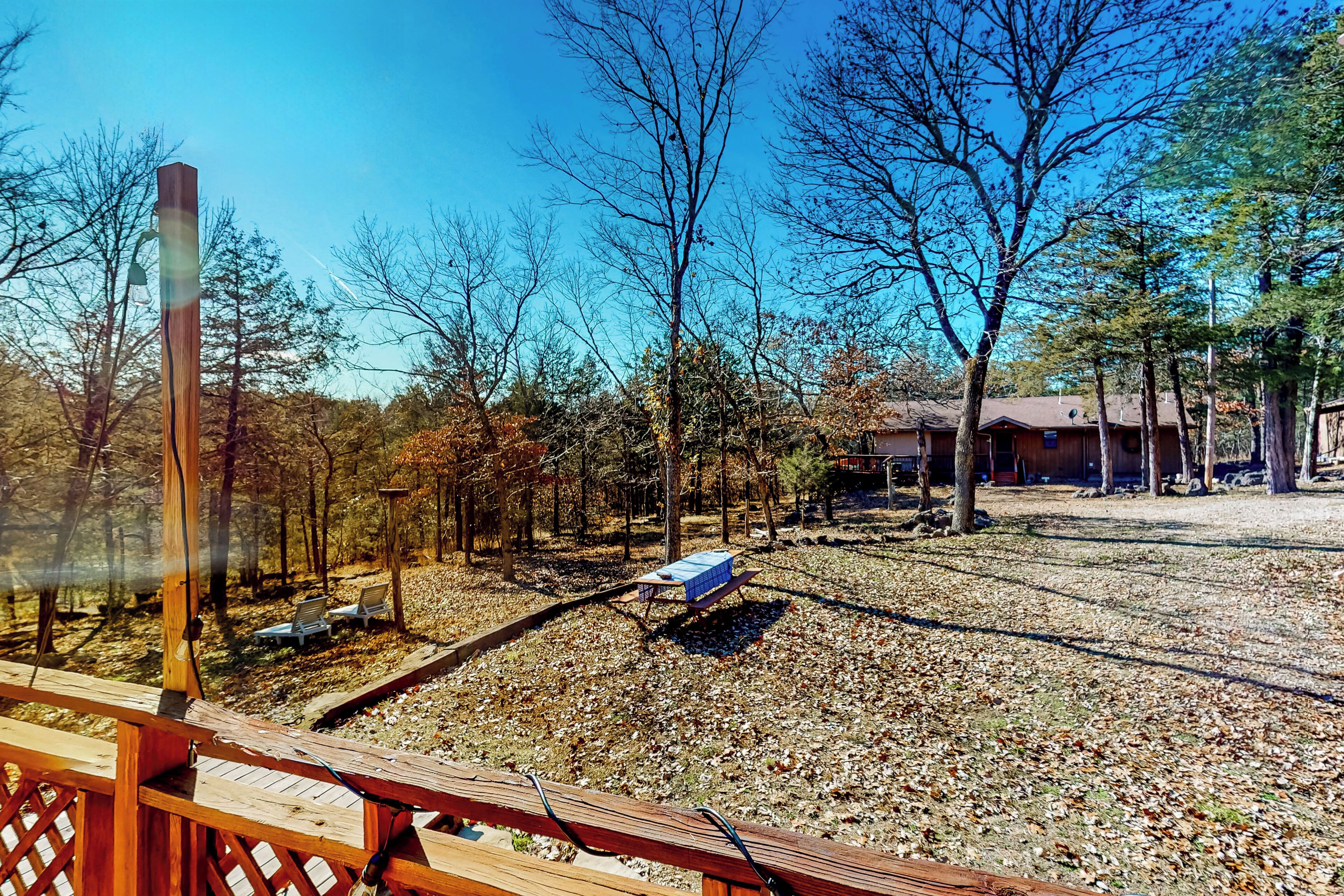 A wooden deck with benches, a picnic table, and a clear view of a wooded area with leaf-covered ground.