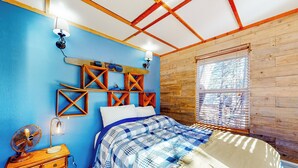 Une chambre avec un mur bleu, des poutres en bois au plafond, un lit recouvert d’une couverture à carreaux bleus et blancs, une commode en bois et une fenêtre avec des stores.