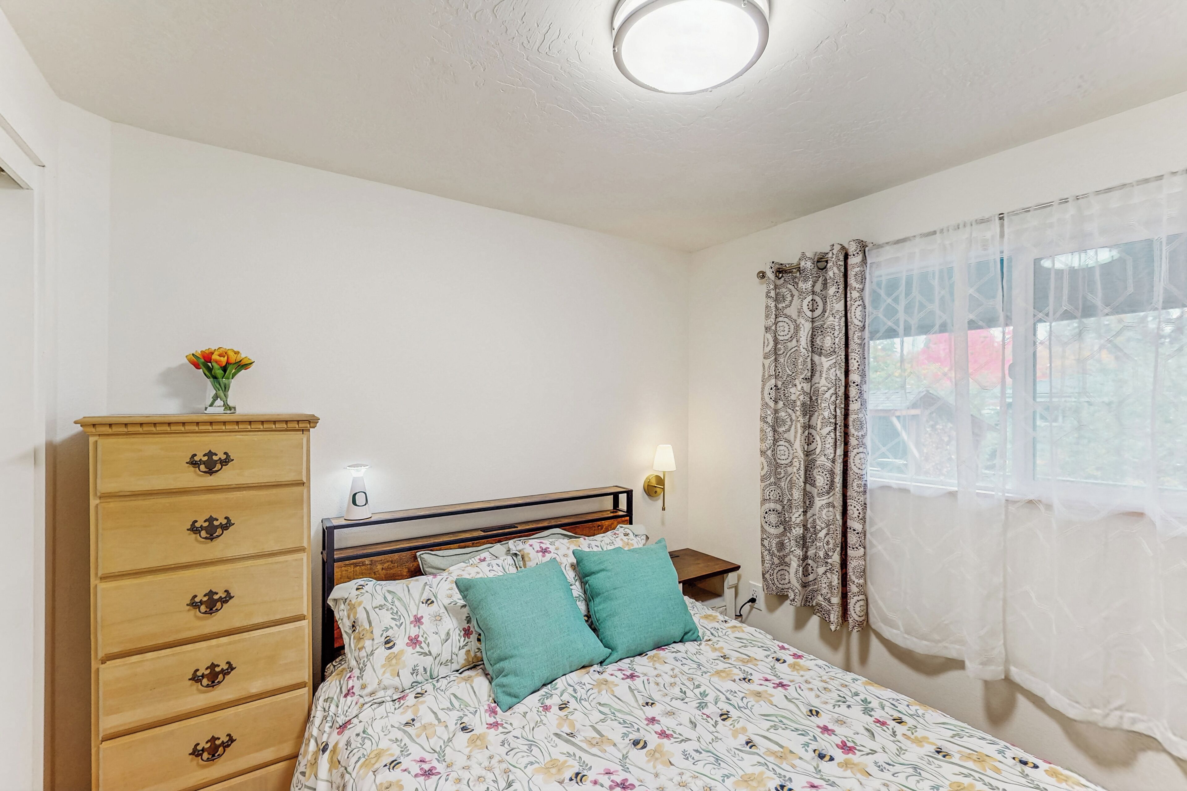 A bedroom with a bed, a wooden dresser, a nightstand, a wall-mounted lamp, and a window with sheer curtains.