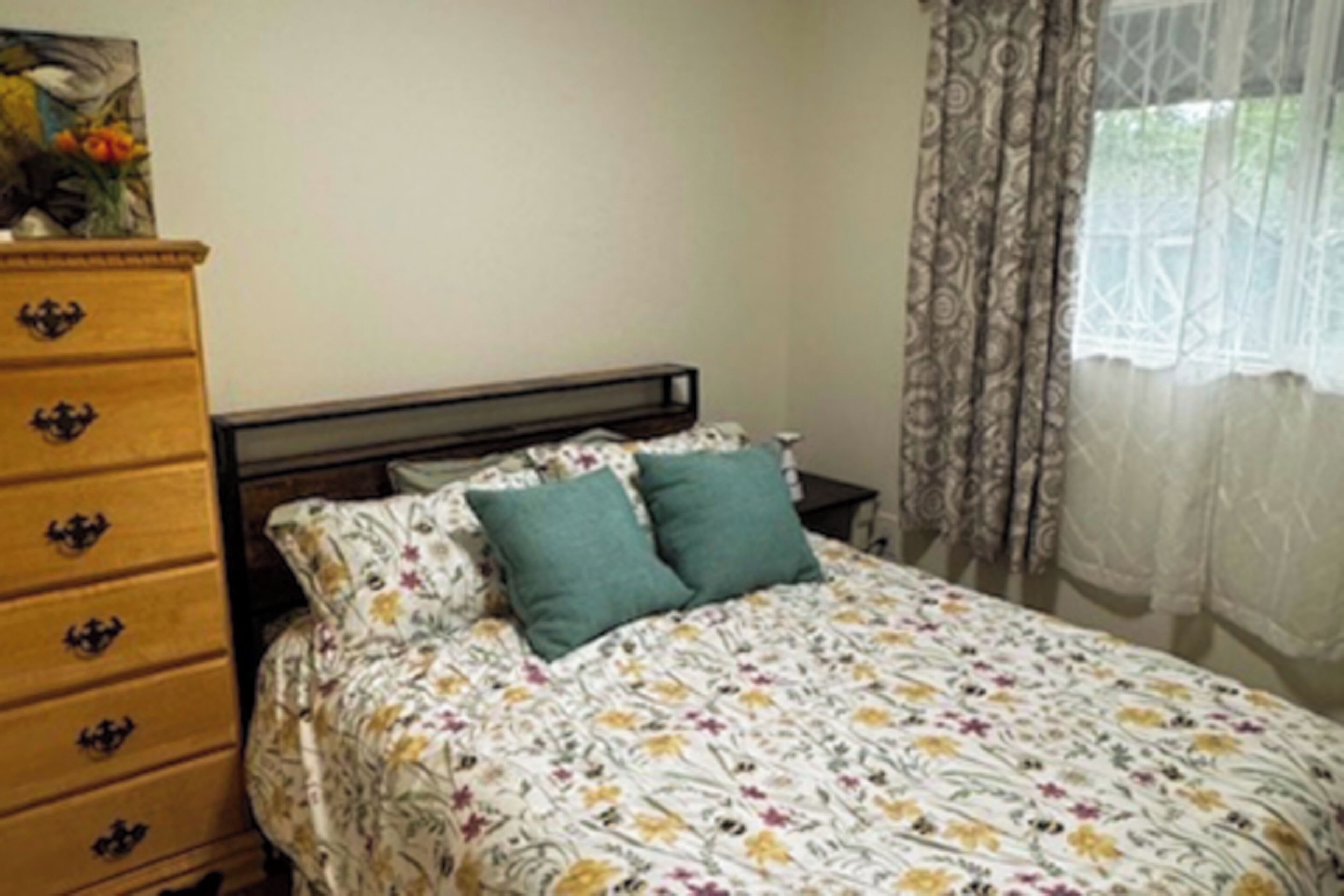 A bedroom with a floral bedspread, a wooden dresser with drawers, and a window with patterned curtains.