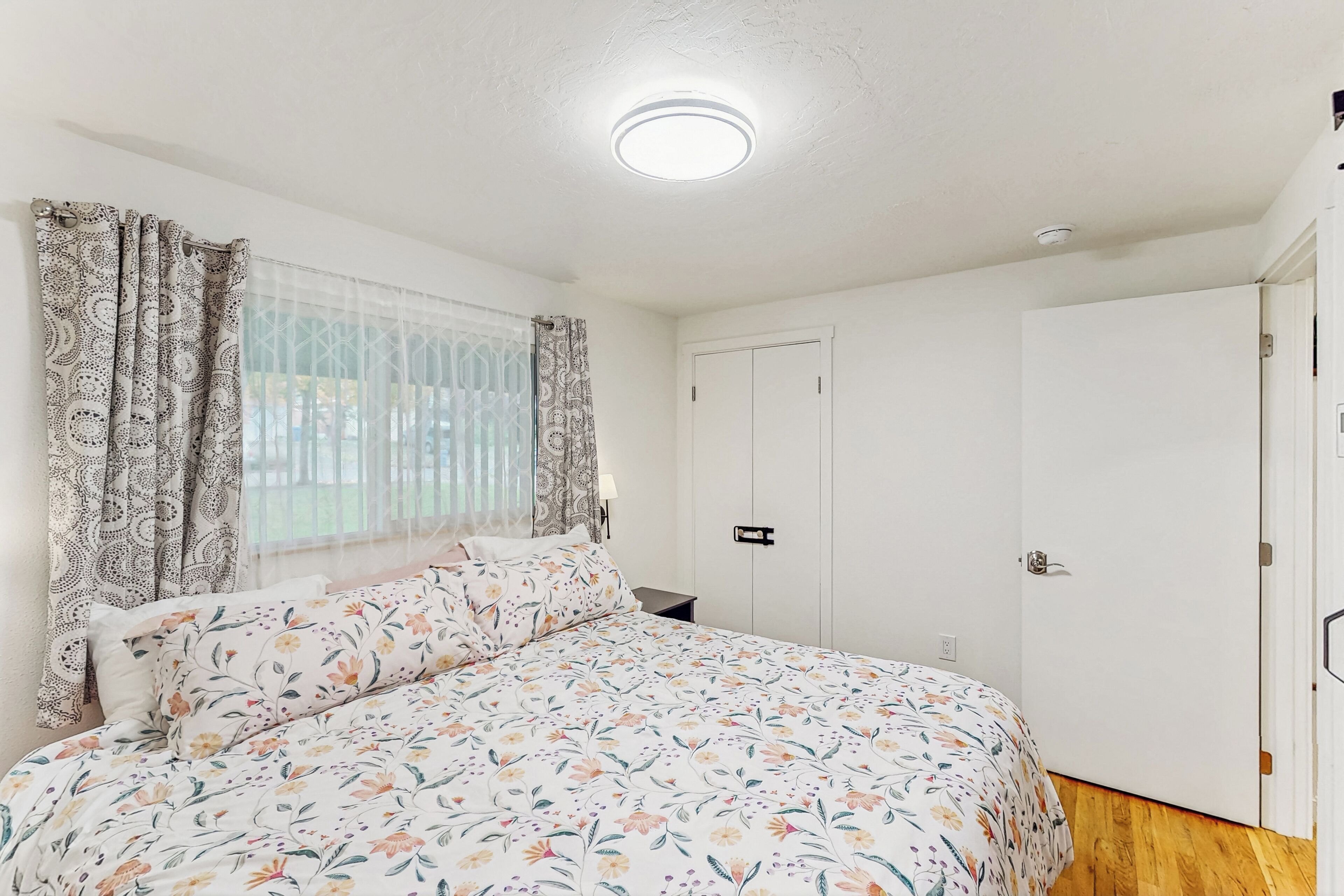 A bedroom with a floral bedspread, a window with patterned curtains, and a white wardrobe.