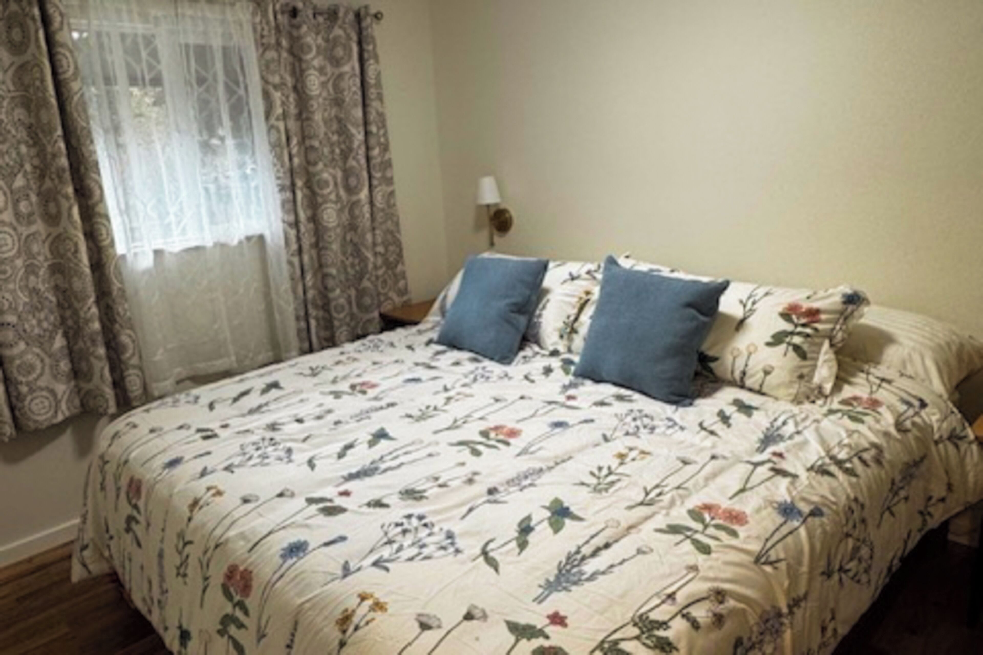 Une chambre à coucher avec une couvre-lit à motifs floraux, deux oreillers, une table de chevet avec une lampe et des rideaux à motifs.