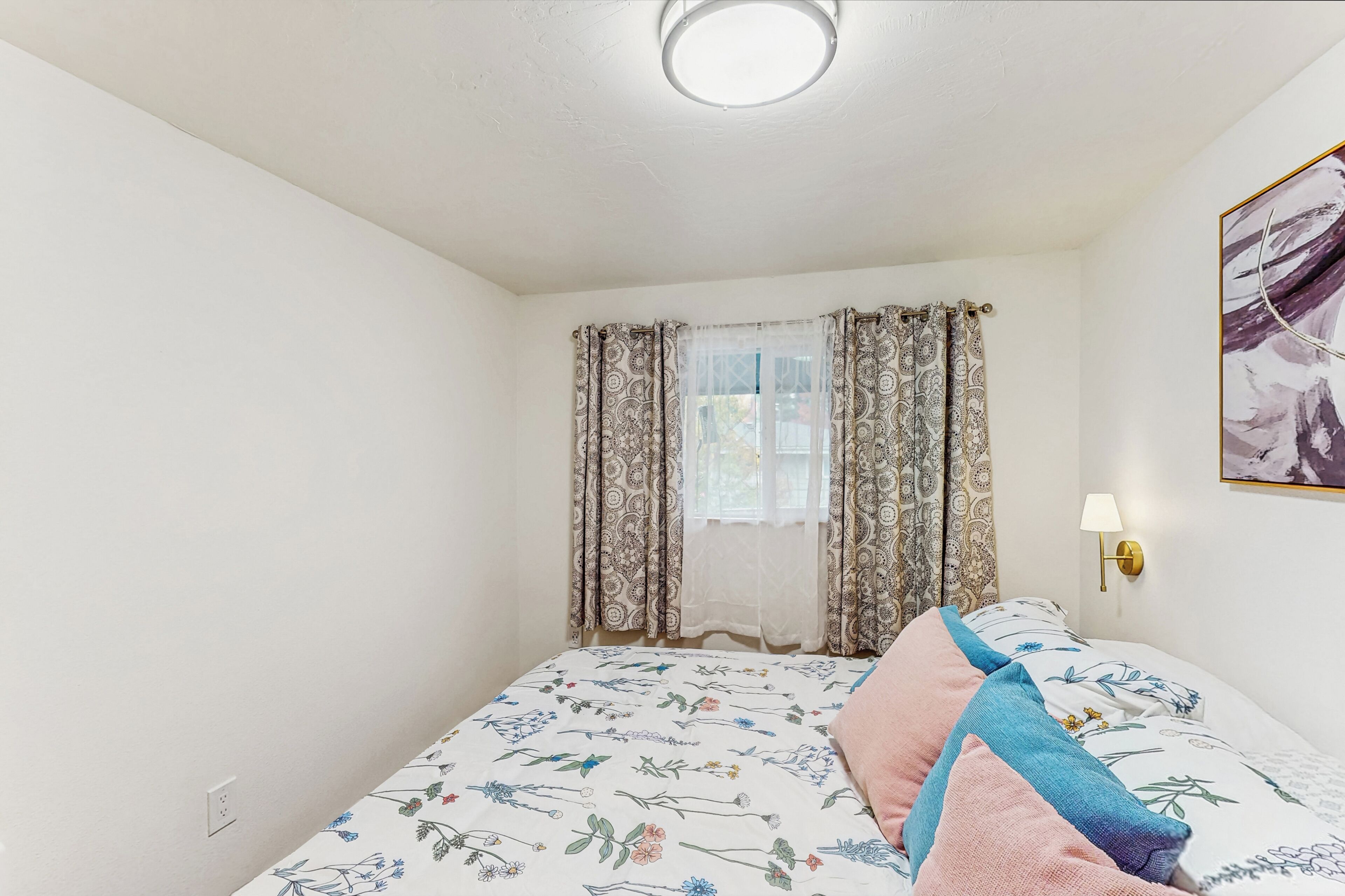 A bedroom with a bed, floral-patterned bedspread, patterned curtains, a wall-mounted lamp, and a framed picture on the wall.