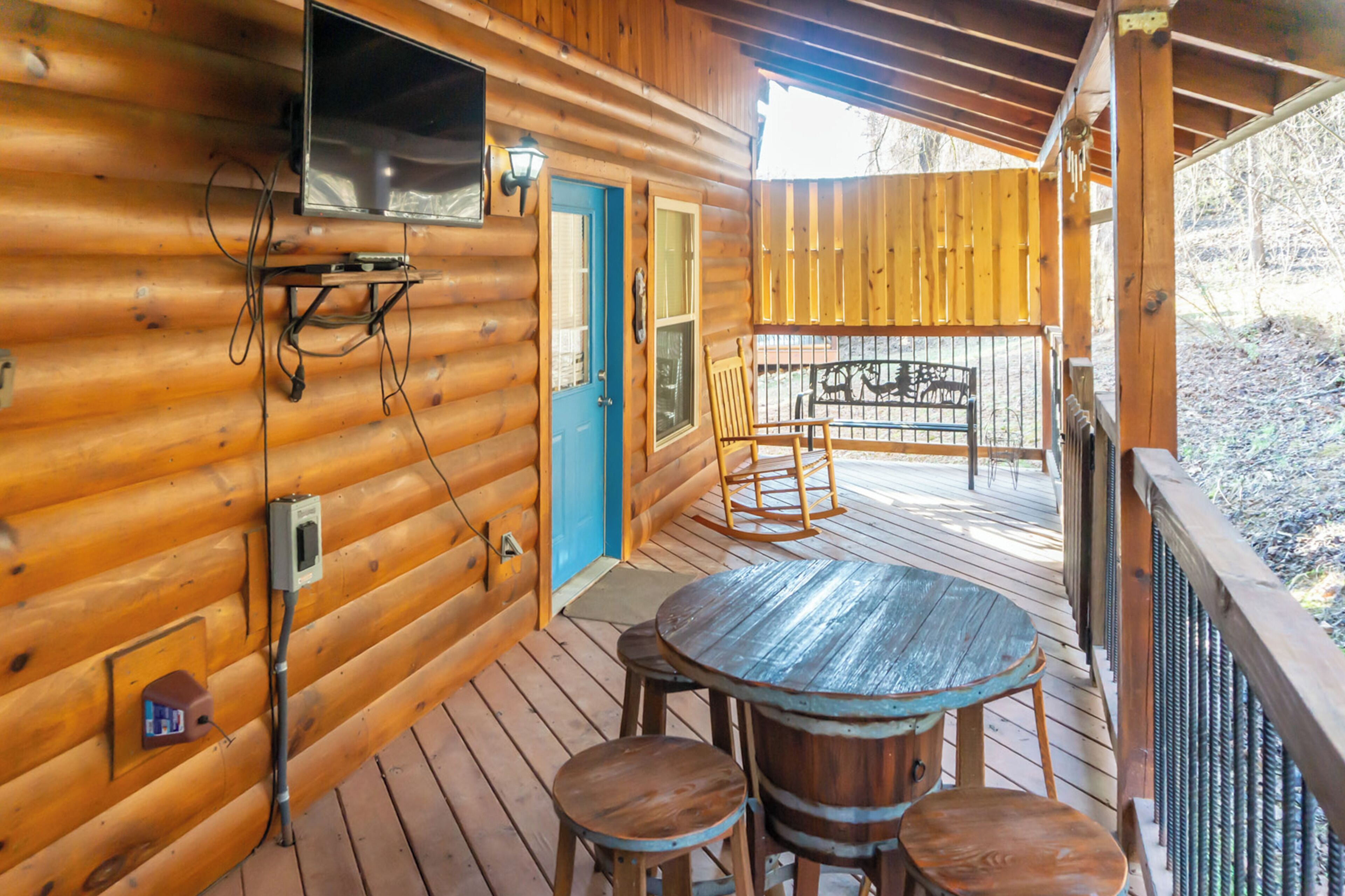 A wooden cabin with a blue door, a TV mounted on the wall, a wooden deck with a table and chairs, and a rocking chair.