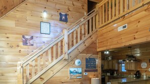 Interior de uma cabana de madeira com escada, paredes rústicas, área de cozinha e diversos adornos nas paredes, incluindo quadros de animais e uma placa de boas-vindas.