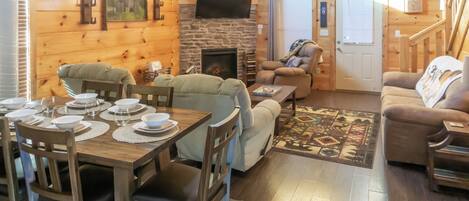 A cozy cabin interior with a dining area, sofas, a fireplace, and a television mounted on the wall.