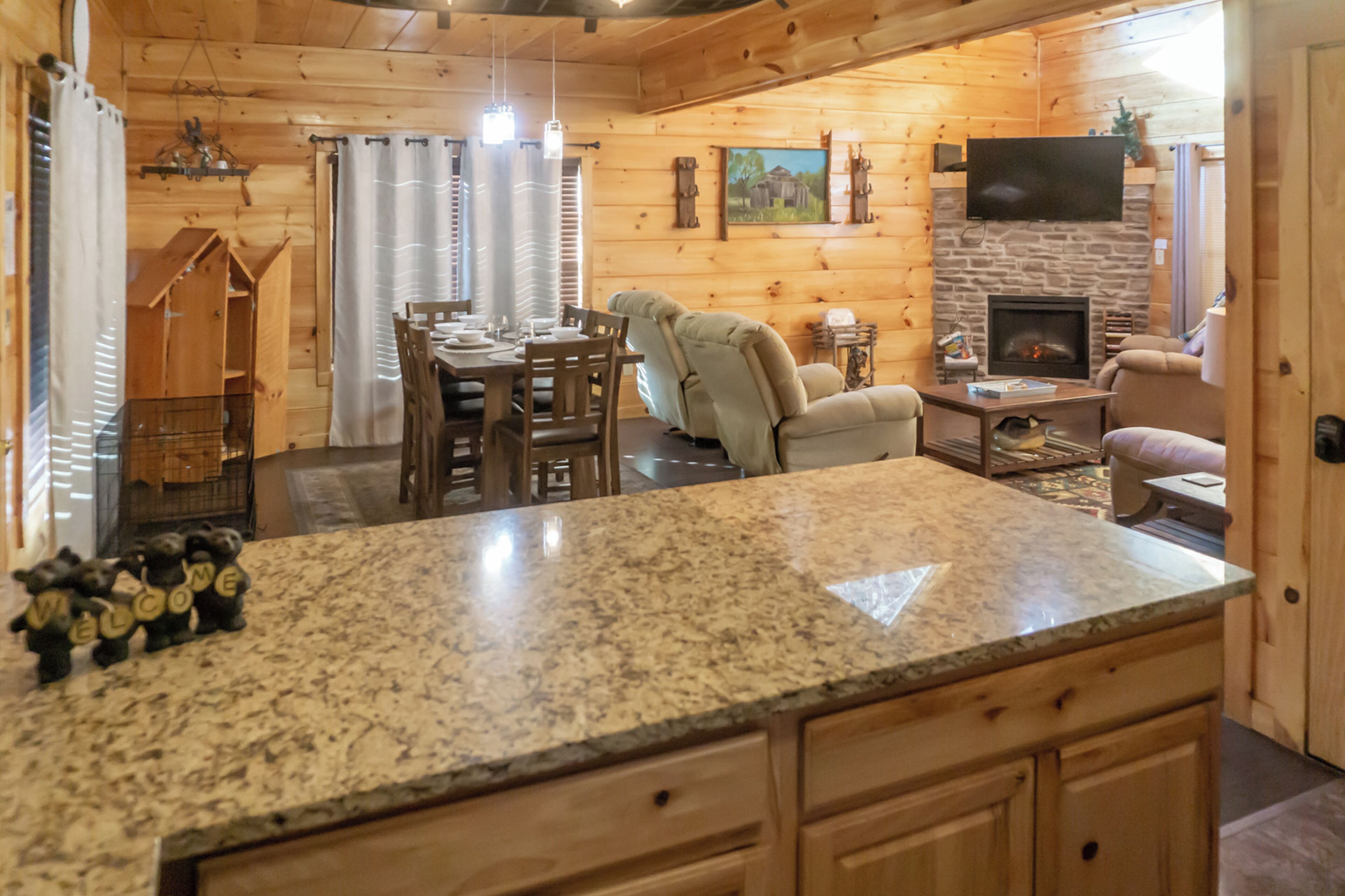 A cozy cabin interior with a stone fireplace, wooden walls, a dining area, and a kitchen with granite countertops.