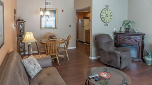 Una sala de estar con un sofá, una mesa de centro de vidrio, un comedor con sillas, una cocina con refrigerador y un reloj en la pared.