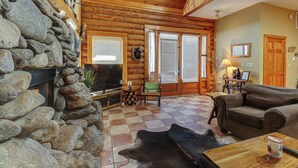 Interior de una cabaña de troncos con pared de piedra, chimenea, televisor, sofá y mesa de comedor.