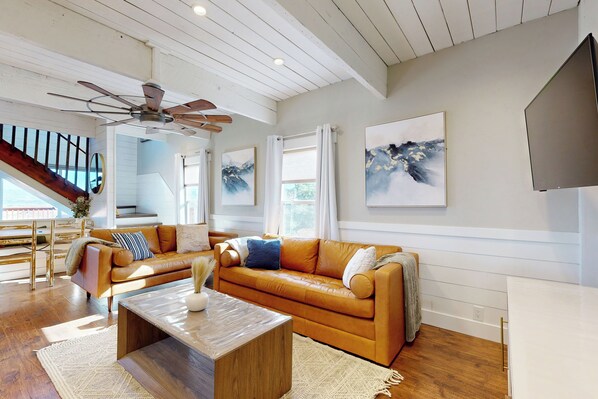 A spacious living room with a large brown leather sofa, a wooden coffee table, and a ceiling fan. There are two large windows with white curtains, a staircase with wooden railings, and a flat-screen TV mounted on the wall. - Private Hot Tub & Sauna! Midway Family Hideaway (Midway)