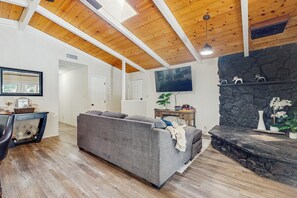 Un salon doté d’un canapé d’angle gris, de poutres apparentes en bois au plafond, d’une cheminée en pierre et d’un téléviseur à écran plat fixé au mur.