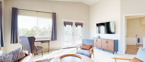 A spacious living room with a large window, a flat-screen TV mounted on the wall, and a dining area visible through the glass doors.