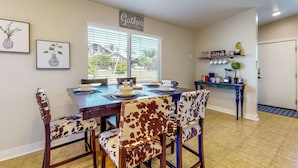 A dining area with a wooden table set for four, surrounded by chairs with a cowhide pattern. A 'Gather' sign is above the window, and a coffee-themed shelf is on the wall. - Two Dog-friendly Home, Close to Wine Country (Fallbrook)