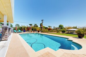 Ein rechteckiger Pool mit Fliesen umgeben, mit einer weißen Umzäunung und einer überdachten Terrasse mit Stühlen und Sonnenschirmen.