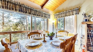 A dining area with a table set for four, surrounded by wooden chairs, large windows with plaid curtains, and a view of trees. - Idyllic Cabin in the Woods: Cozy Crestline Retreat (Crestline)