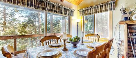 A dining area with a table set for four, surrounded by wooden chairs, large windows with plaid curtains, and a view of trees.