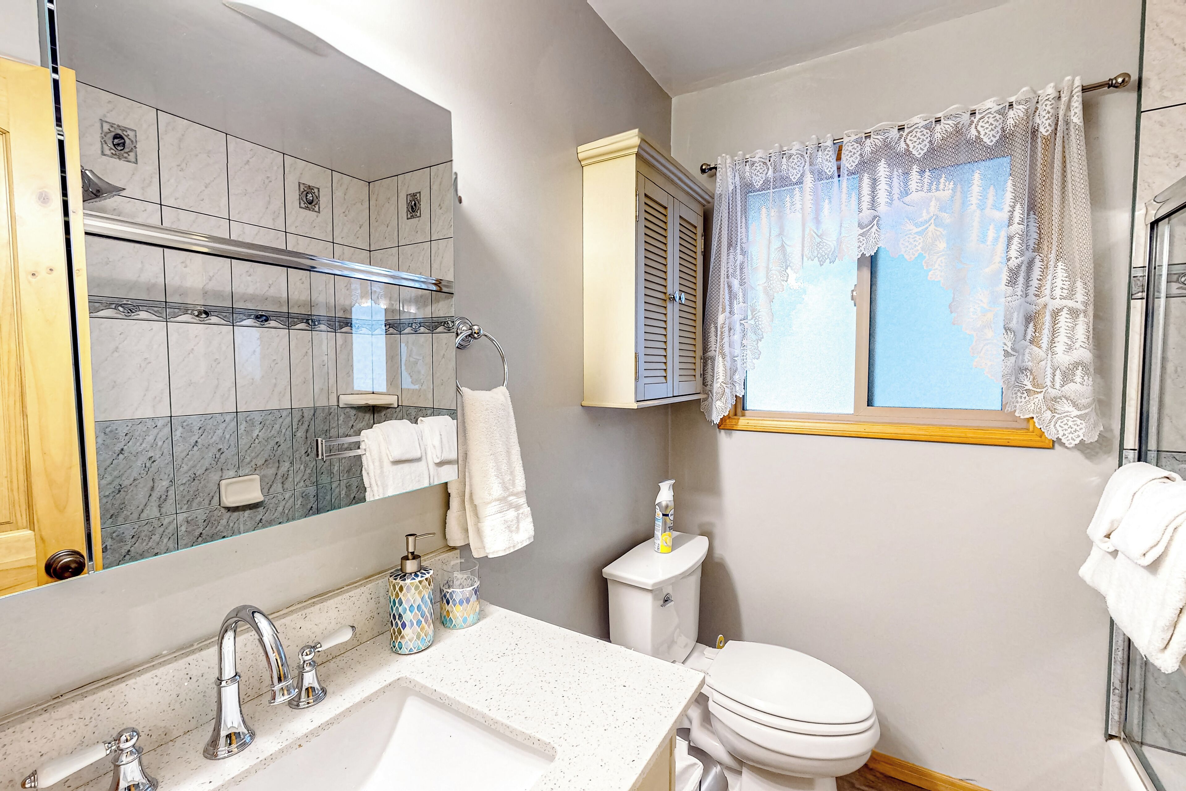 A bathroom with a large mirror, a sink with two faucets, a toilet, and a window with lace curtains.