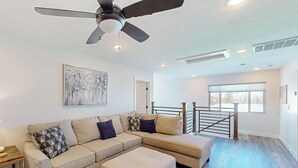 A modern living room with a sectional sofa, ottoman, ceiling fan, artwork, and a view through a large window. - Crooked River 10 A - Entire Home! (Brian Head)