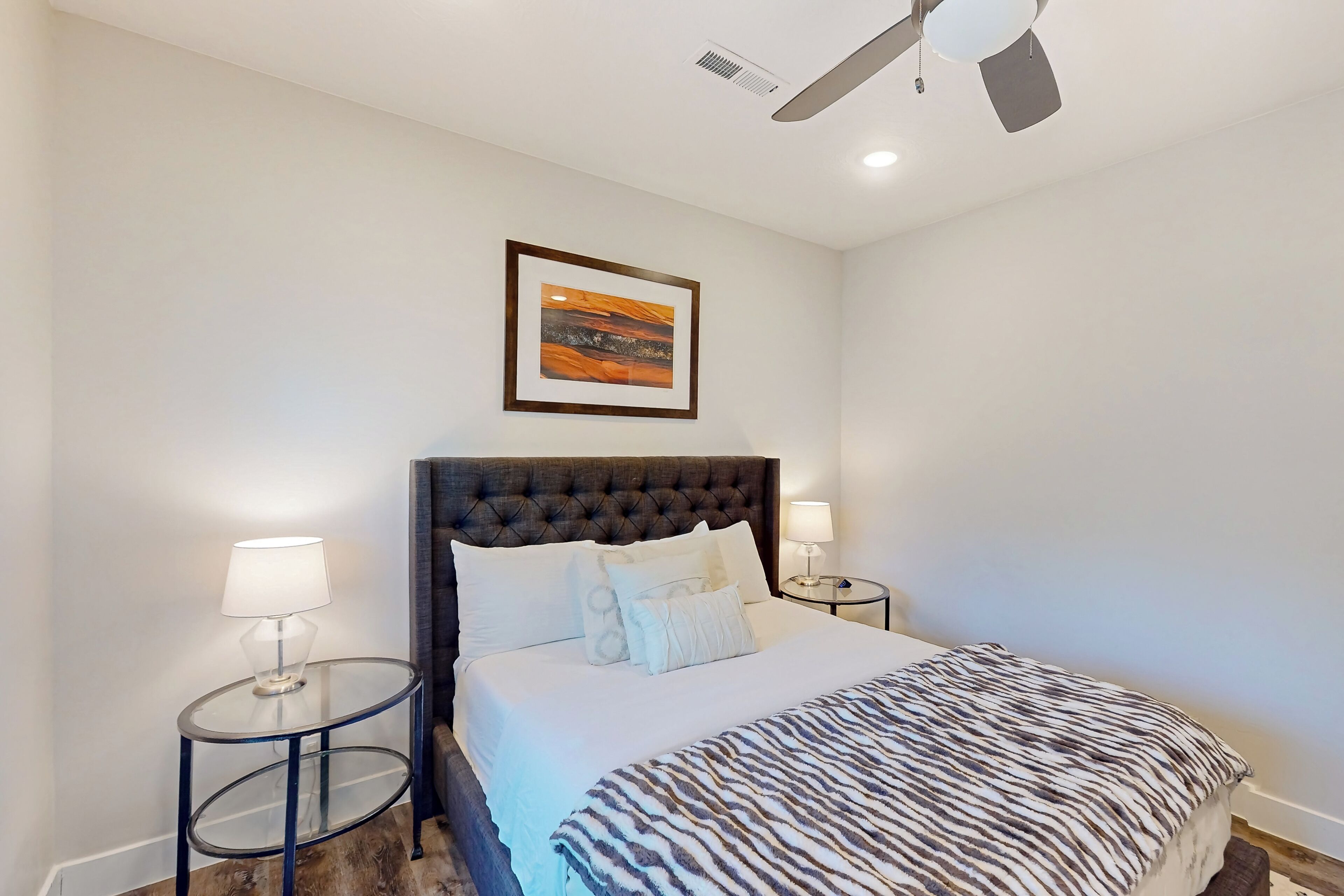 A neatly arranged bedroom with a tufted headboard, a zebra-patterned bedspread, two bedside lamps, and a framed artwork on the wall.