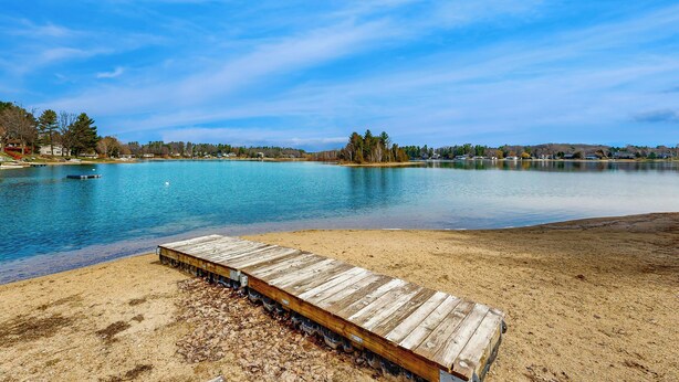 A lakeside view with a wooden dock extending into calm water, surrounded by trees and distant buildings.