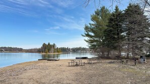 A lakeside picnic area with benches and picnic tables, surrounded by trees and a clear sky.