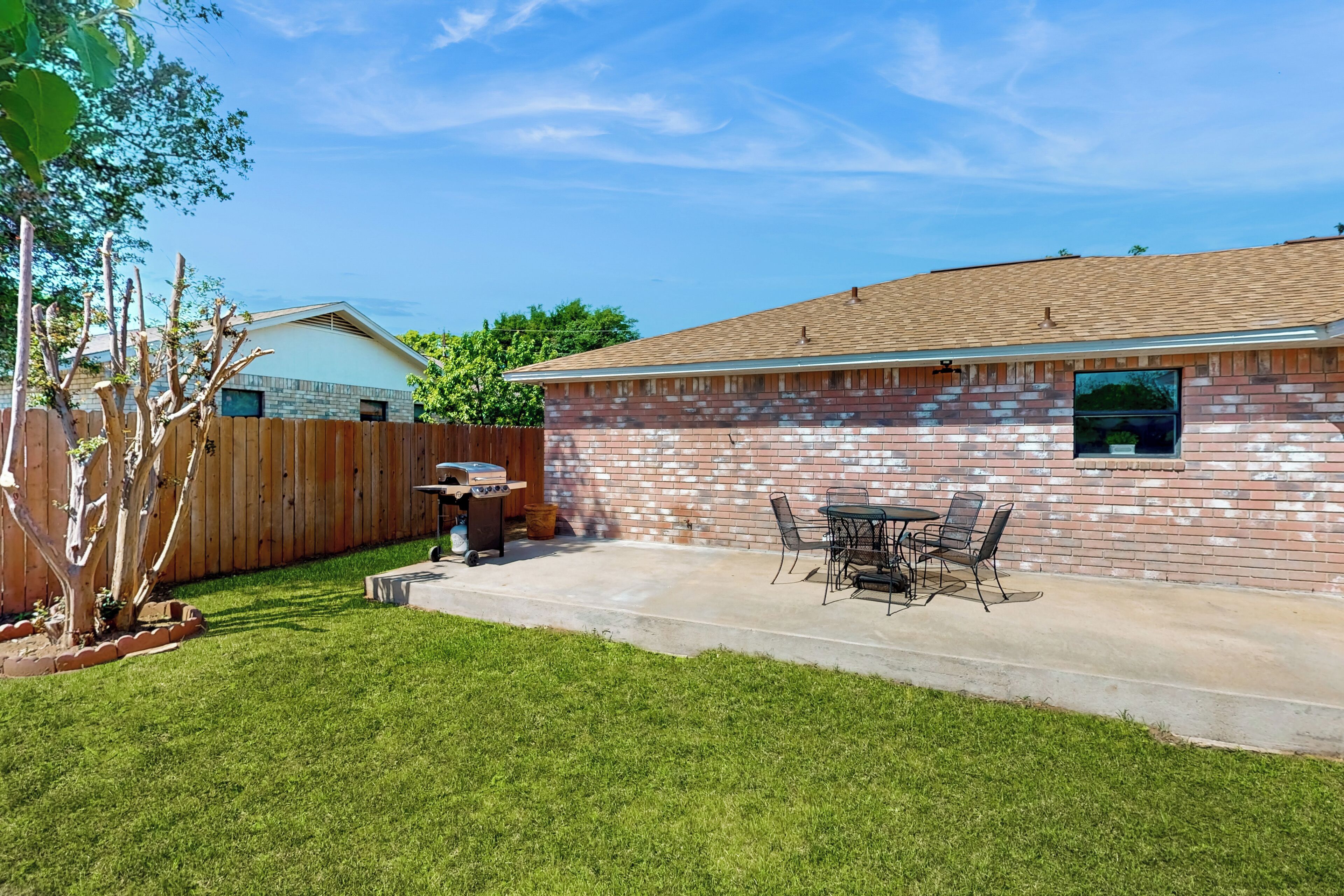 A backyard with a brick patio, a wooden fence, a barbecue grill, and a dining table with chairs.
