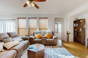 A living room with a sofa, armchair, and coffee table. There are two windows with blinds, a ceiling fan, and a bookshelf. - Inviting Home w/ Enlosed Yard & Gas Grill (Round Rock)