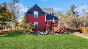 A red house with a dark roof, a front porch with chairs, and a small garden. - 4BR House w/ Kitchen, Jetted Tub, Wifi + gas Grill (Orrs Island)