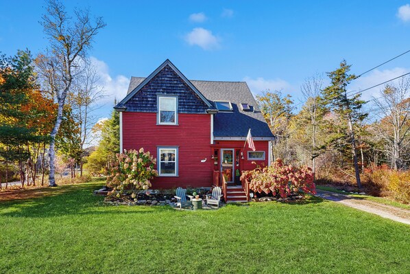 Une maison rouge avec un toit foncé, une véranda à l’avant avec des chaises et un petit jardin.
