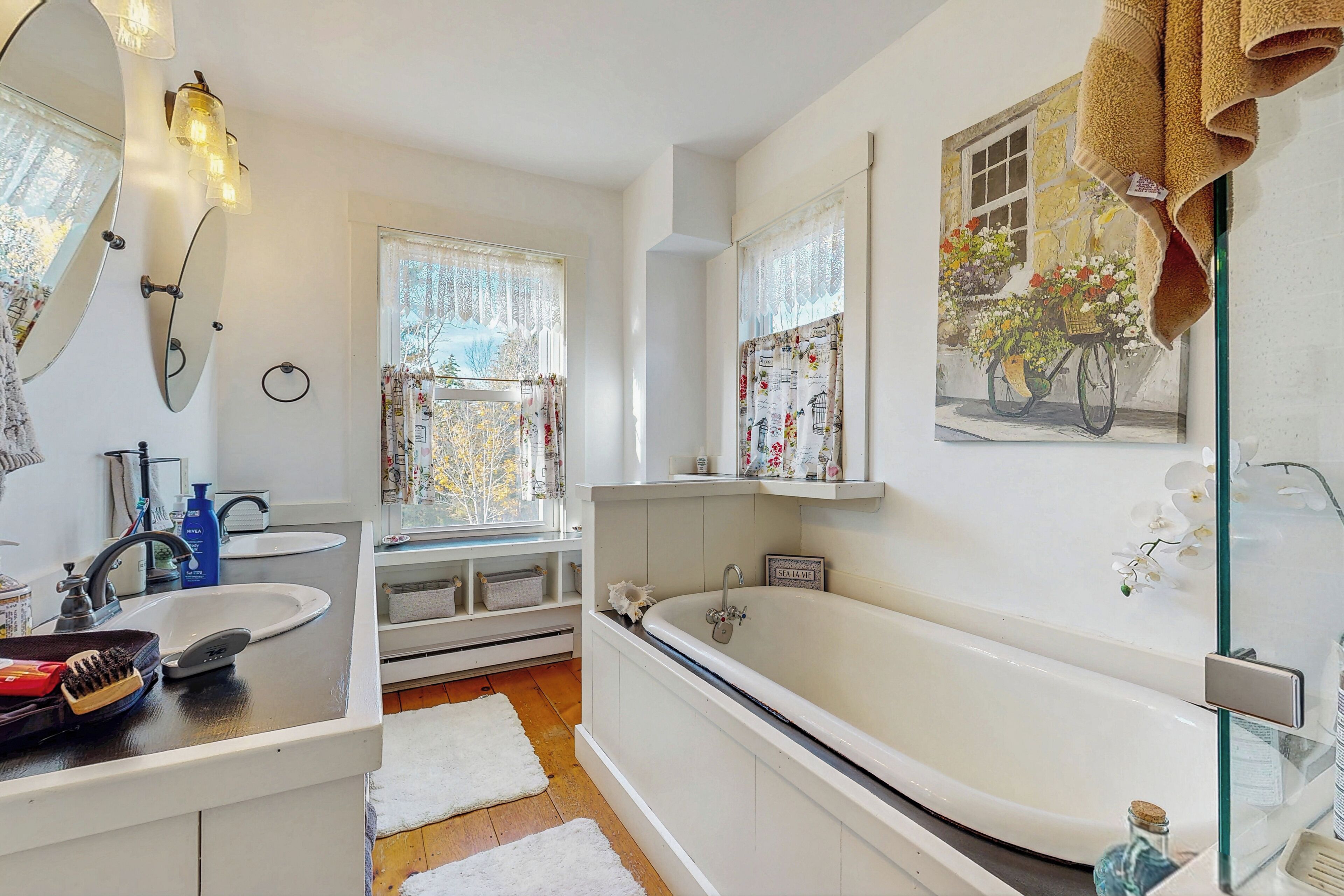 Une salle de bain moderne avec une baignoire, un lavabo et un miroir. On y trouve des fenêtres avec des rideaux, un porte-serviettes et une étagère avec des articles de toilette.
