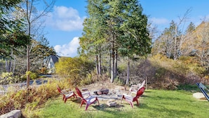 A circular seating area with a fire pit surrounded by red chairs, set in a grassy yard with trees and a house in the background. - 4BR House w/ Kitchen, Jetted Tub, Wifi + gas Grill (Orrs Island)