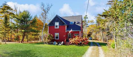 A red house with a front porch, a lawn, and trees with autumn leaves.