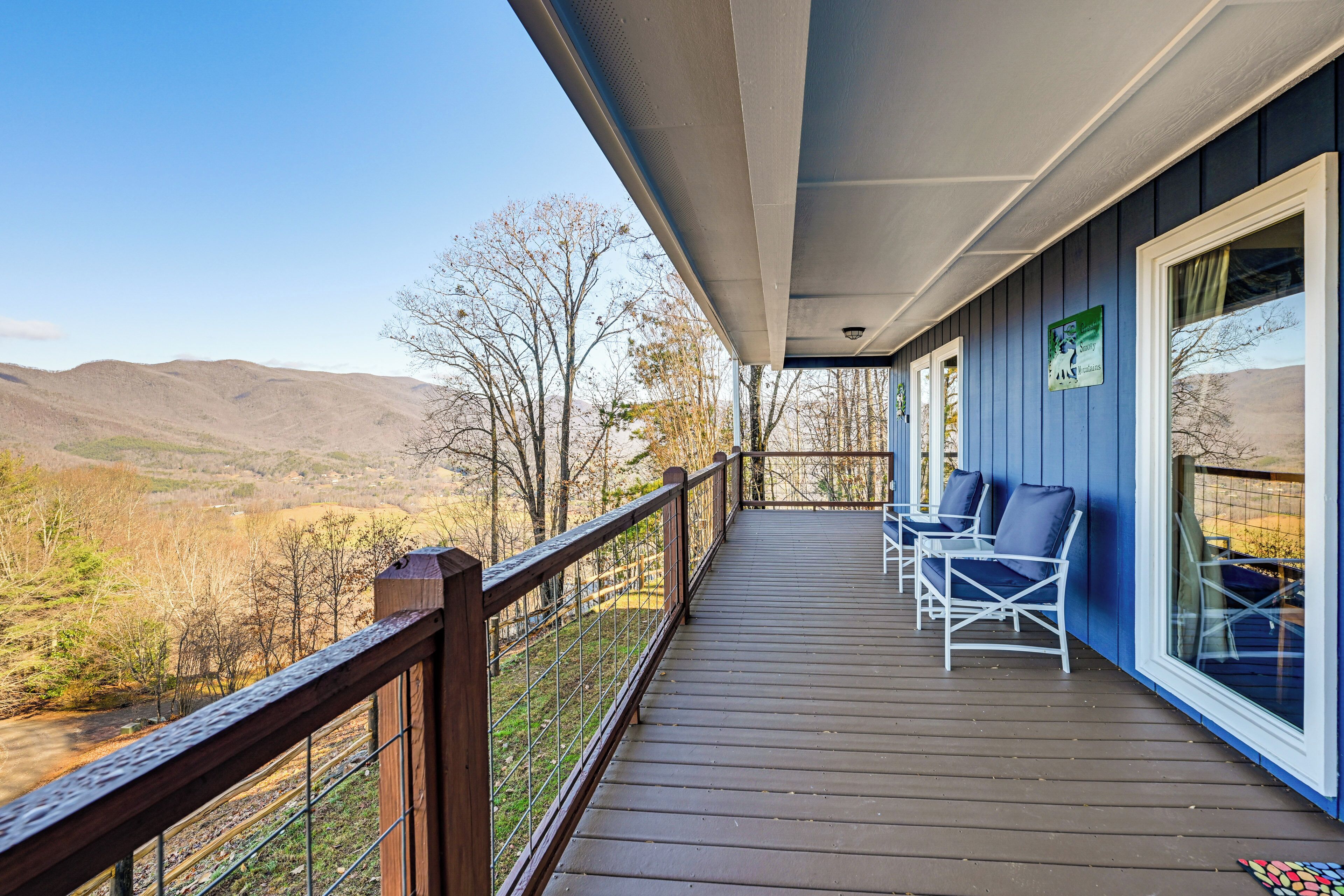 A wooden deck with a railing, two white chairs, and a view of mountains and trees.