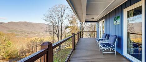 A wooden deck with a railing, two white chairs, and a view of mountains and trees.