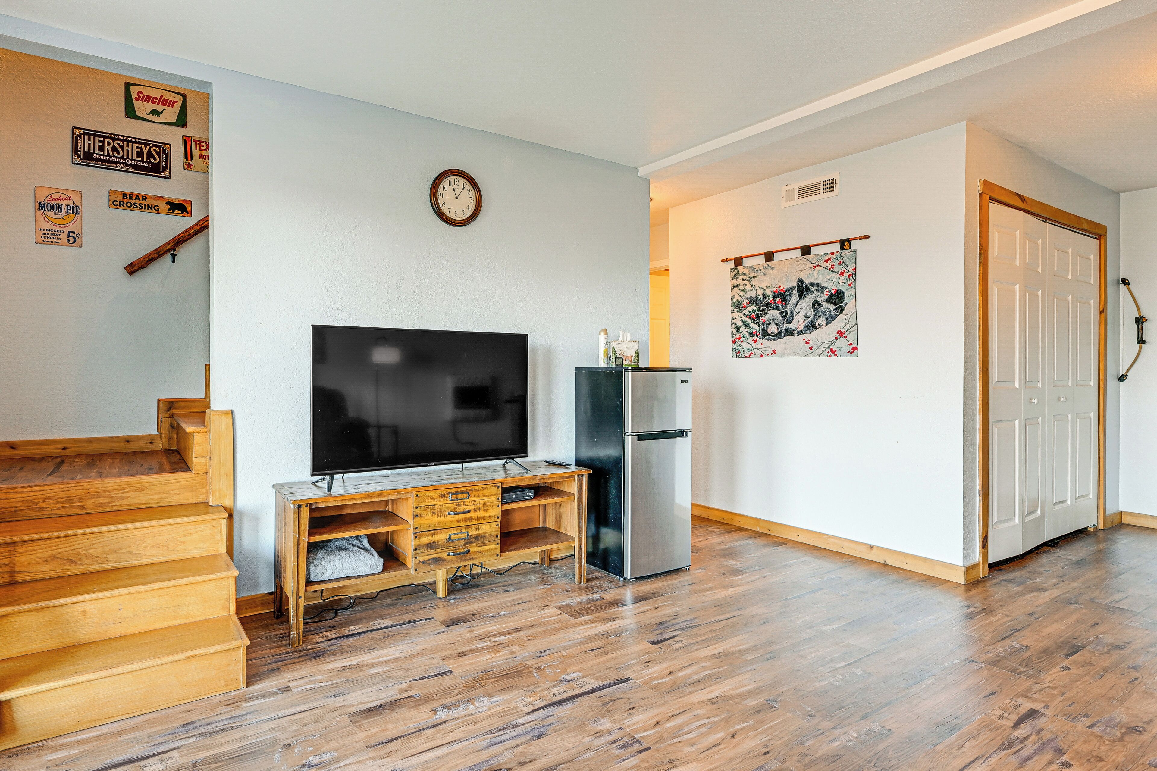 Een woonkamer met een houten trap, een flatscreen televisie, een koelkast en een wandklok.